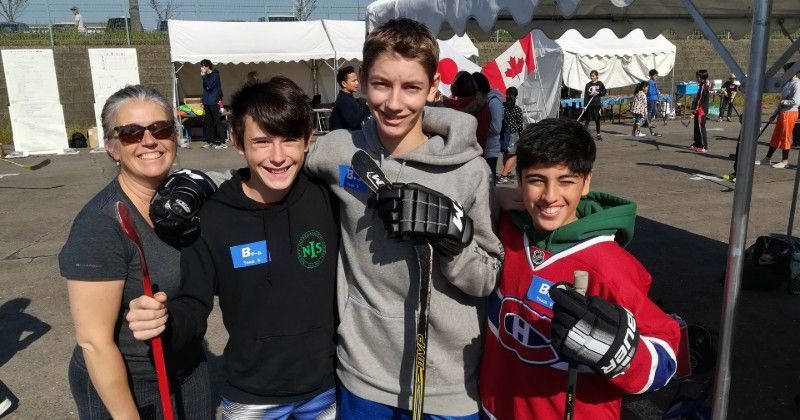 A woman and three boys smile, holding hockey sticks at an outdoor event; a Canadian flag in the background.