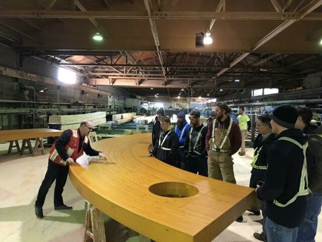 Group observes a wooden structure in a factory; a man in a vest points, explaining details.