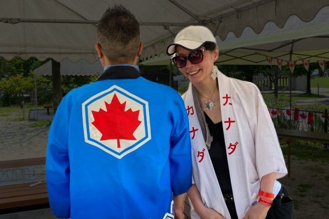 Two people wearing Canada-themed attire. One wears a jacket with a maple leaf, the other a Canada banner. Outdoors.