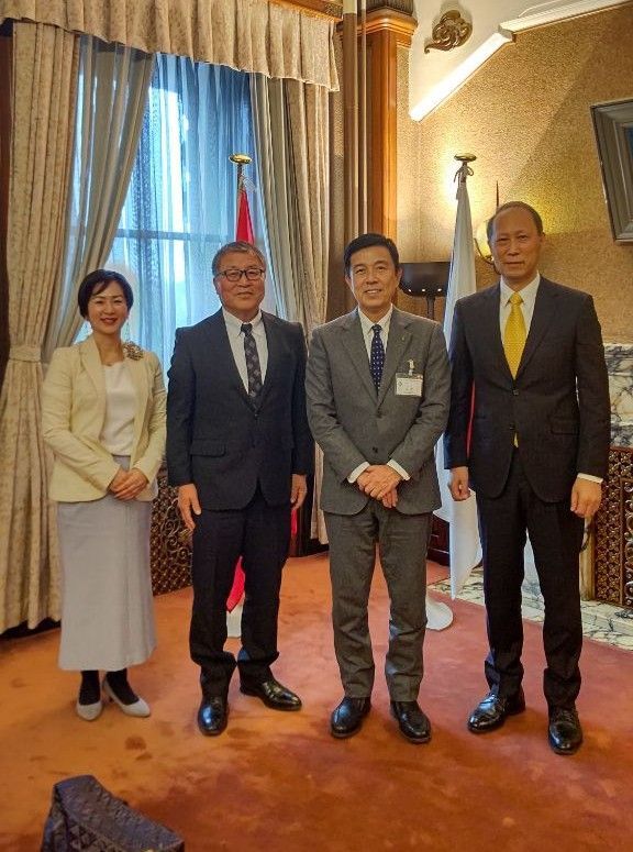 Four people, two men and two women, standing in a room with flags, a window, and a patterned wall.