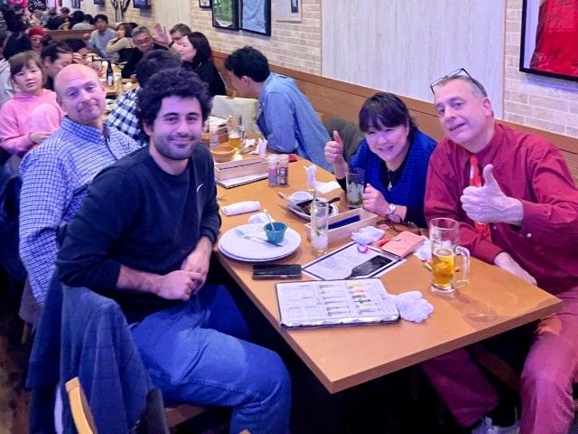 People at a restaurant table, smiling, with drinks and menu. One person gives a thumbs-up.