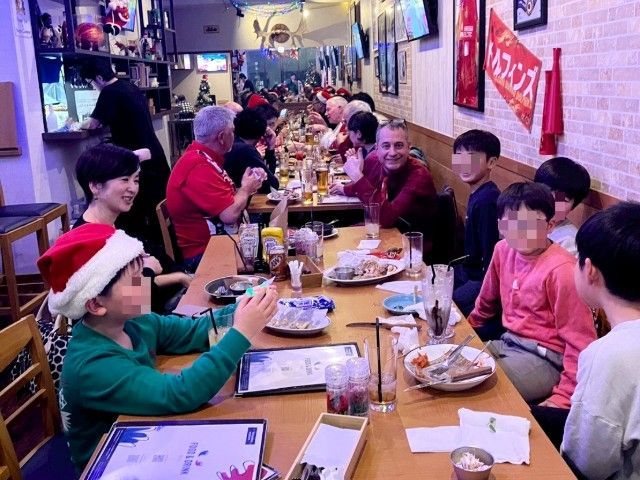 People gathered at a restaurant table, some wearing Santa hats. Colorful decor, food, drinks visible.