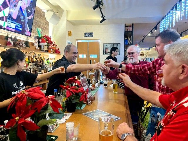 People toasting at a bar with drinks. Bartender and patrons raise glasses. Christmas decorations visible.