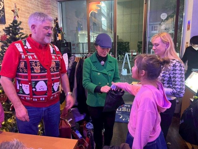 People at an event. A woman in green hands something to a child. A man wears a Christmas vest.
