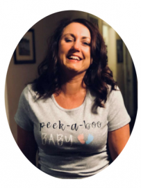 Smiling woman in a gray “peek-a-boo baby” T-shirt standing indoors.