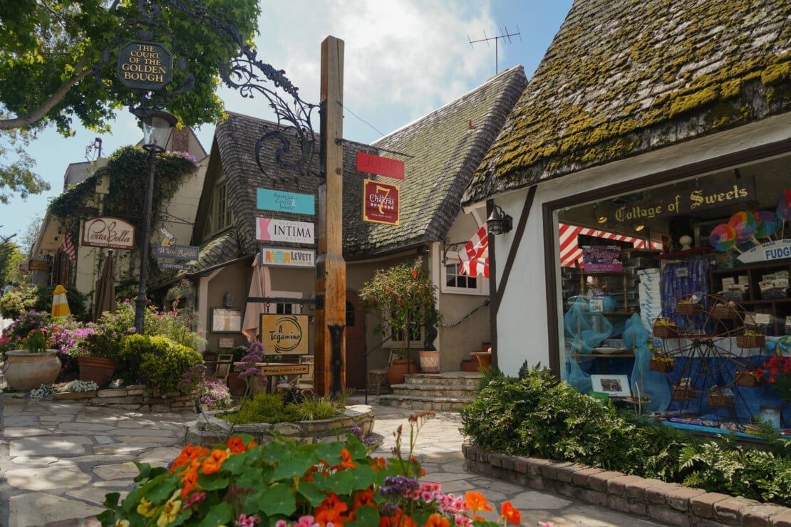 Carmel Village Charming European-style village street in Carmel-by-the-Sea, California, featuring boutique shops, flower-filled walkways, and moss-covered rooftops under a bright sky.