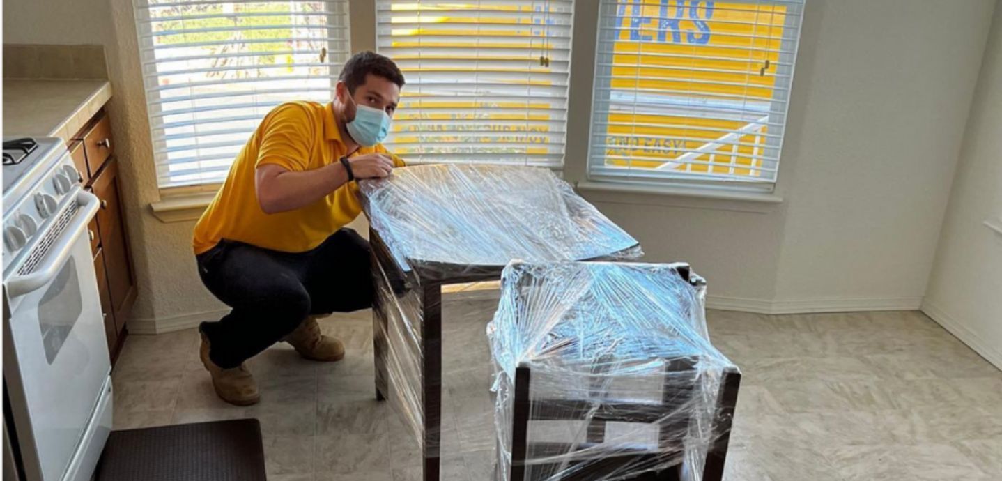 A man wearing a mask is kneeling down next to a table in a kitchen.