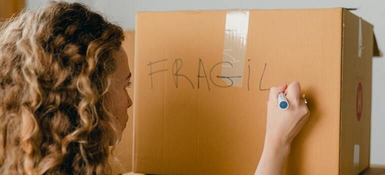 A woman is writing the word fragile on a cardboard box.