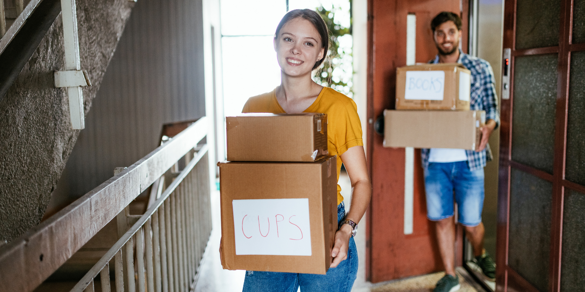 Smiling young couple carrying labeled moving boxes in an apartment building hallway. 