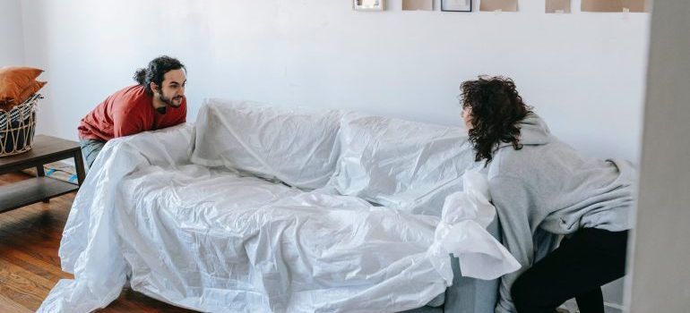 A couple lifting a sofa covered in protective material.