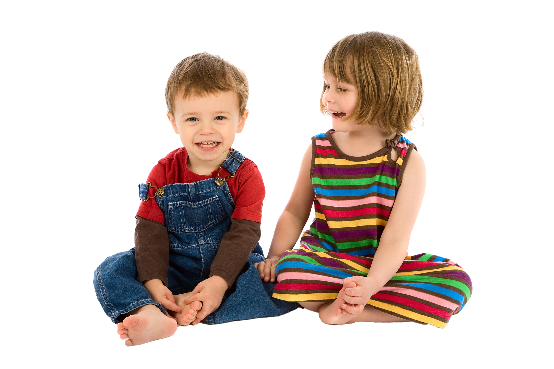 Two young children sitting close together, smiling at the camera. One wears a striped dress; the other has overalls. Two young children sitting close together, smiling at the camera. One wears a striped dress; the other has overalls.