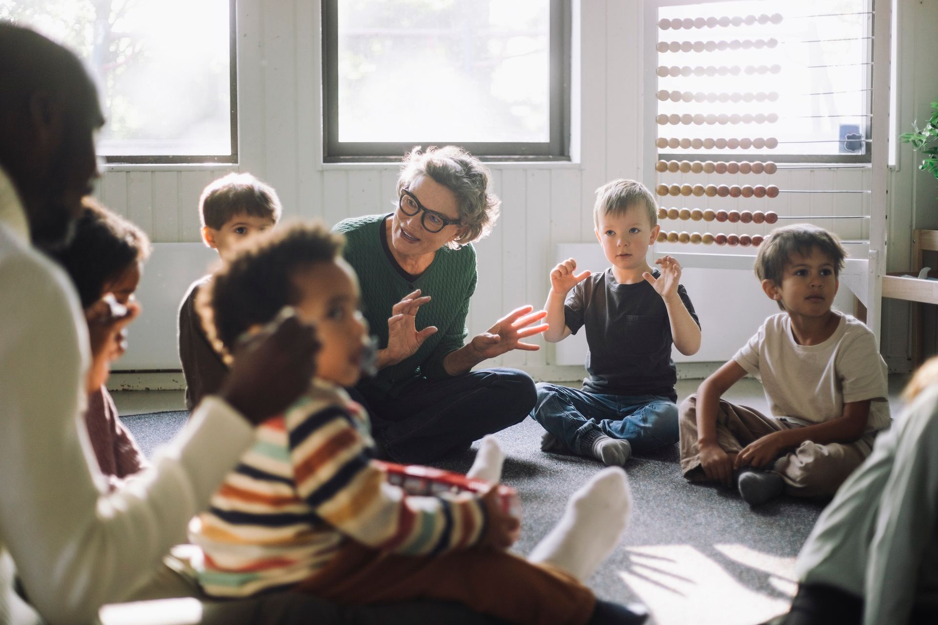 Teacher and children seated on floor in a classroom, one child holding an object, sunlight streaming in.