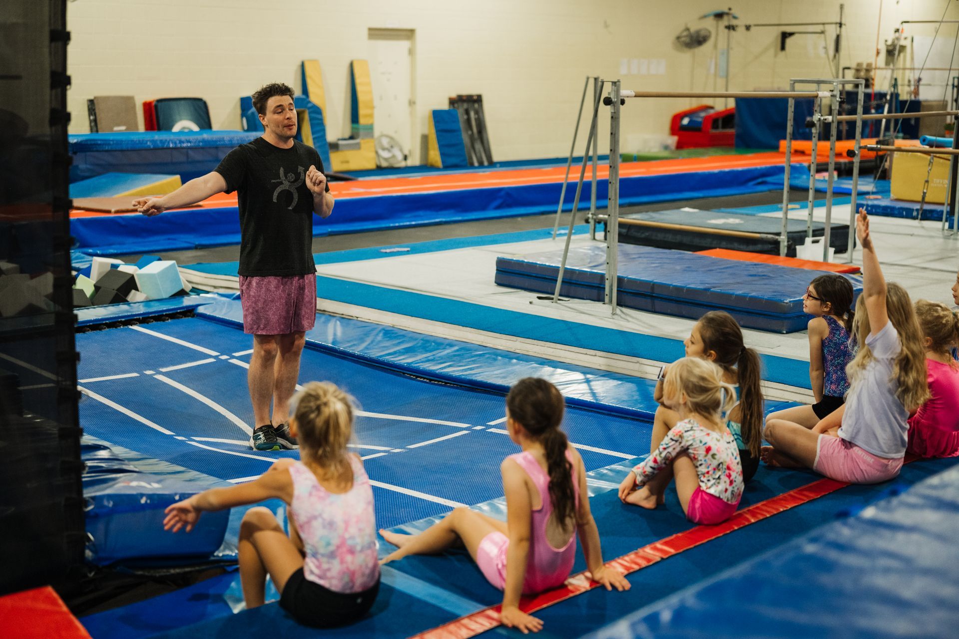 A group of young girls are sitting on the floor in a gym while a man talks to them.