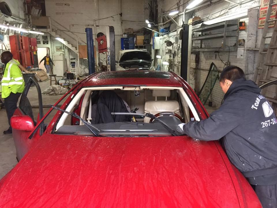 A person is installing a windshield on a red car inside a garage. Another person stands by wearing a safety vest.