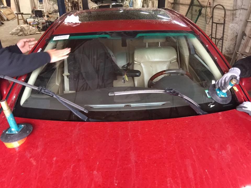 Red car with windshield replacement in progress, suction cups holding glass, indoor shop.