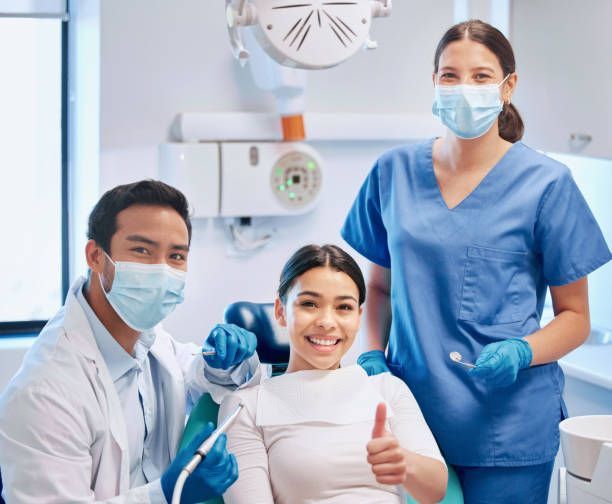 Dentist and assistant with patient in dental chair, all wearing masks, patient giving thumbs up.