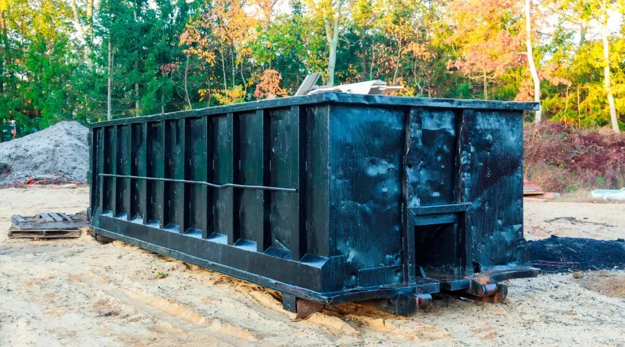 Black dumpster on a dirt surface in a construction area, surrounded by trees and a light sky.