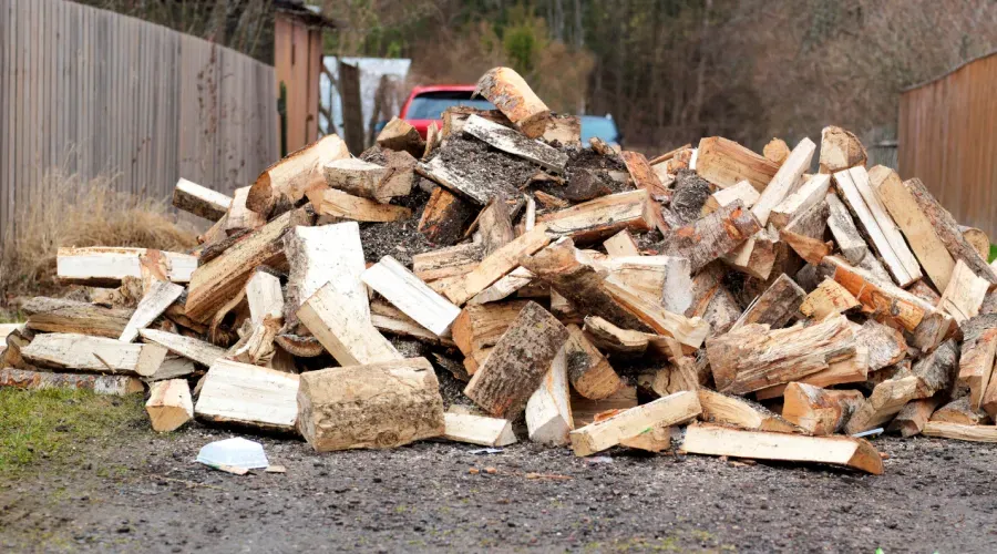 Pile of split firewood on a dirt surface near a wooden fence, a red pickup truck is visible in the background.