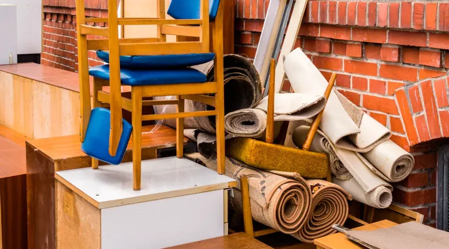 Pile of old furniture and rolled-up rugs against a brick wall. A wooden chair with blue cushions sits on top.