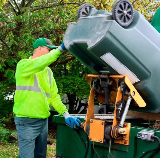 Garbage truck worker in neon green emptying a green trash bin into a mechanical lift. Outdoors, sunny.