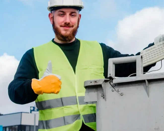 Construction worker with a beard and safety gear giving a thumbs-up. He is next to a gray bin against a blue sky.