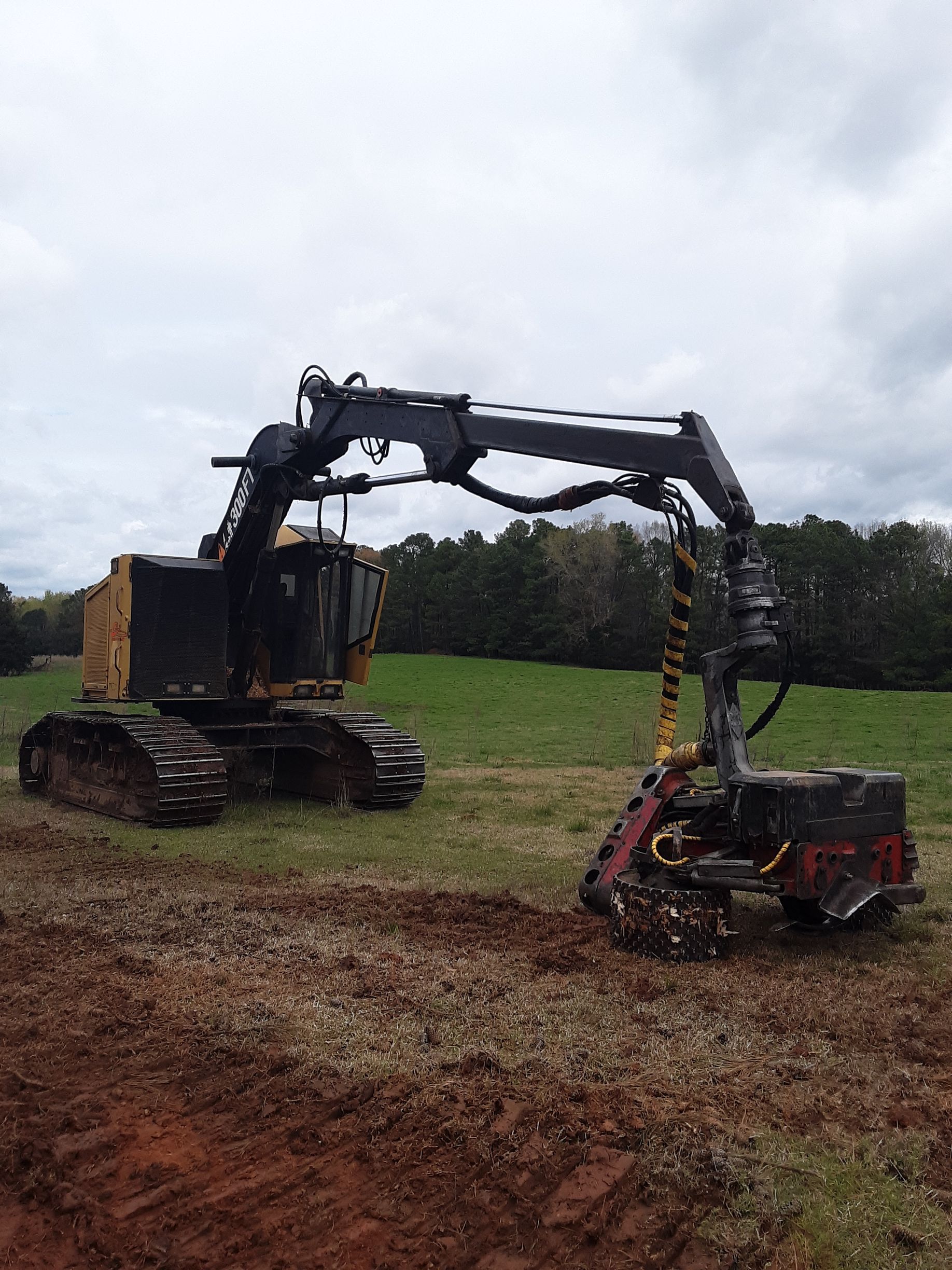 Heavy forestry machine on tracks, cutting trees in a field with a cloudy sky in the background.