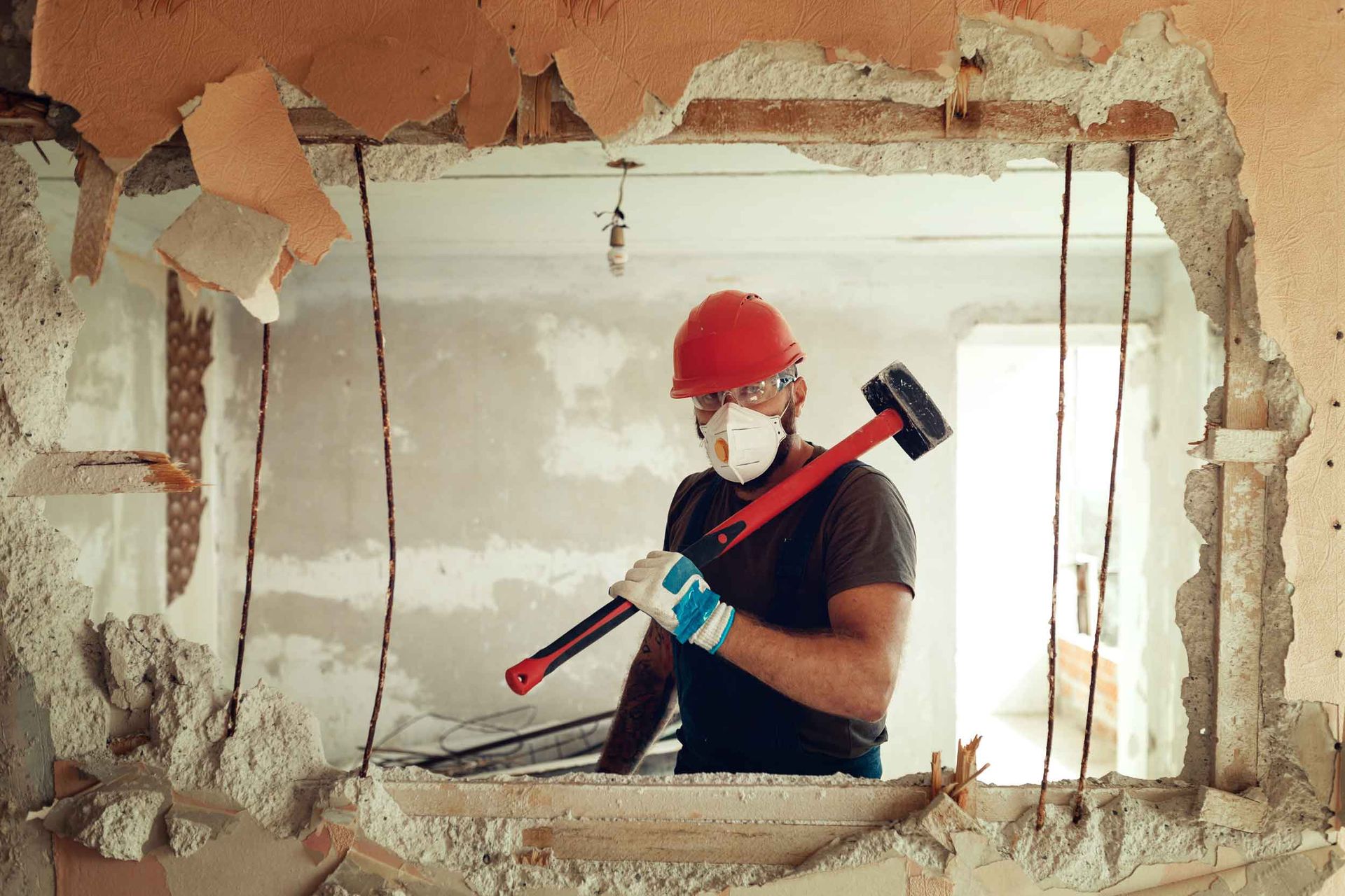 A worker performing demolition by breaking an interior wall with a sledgehammer in an indoor area