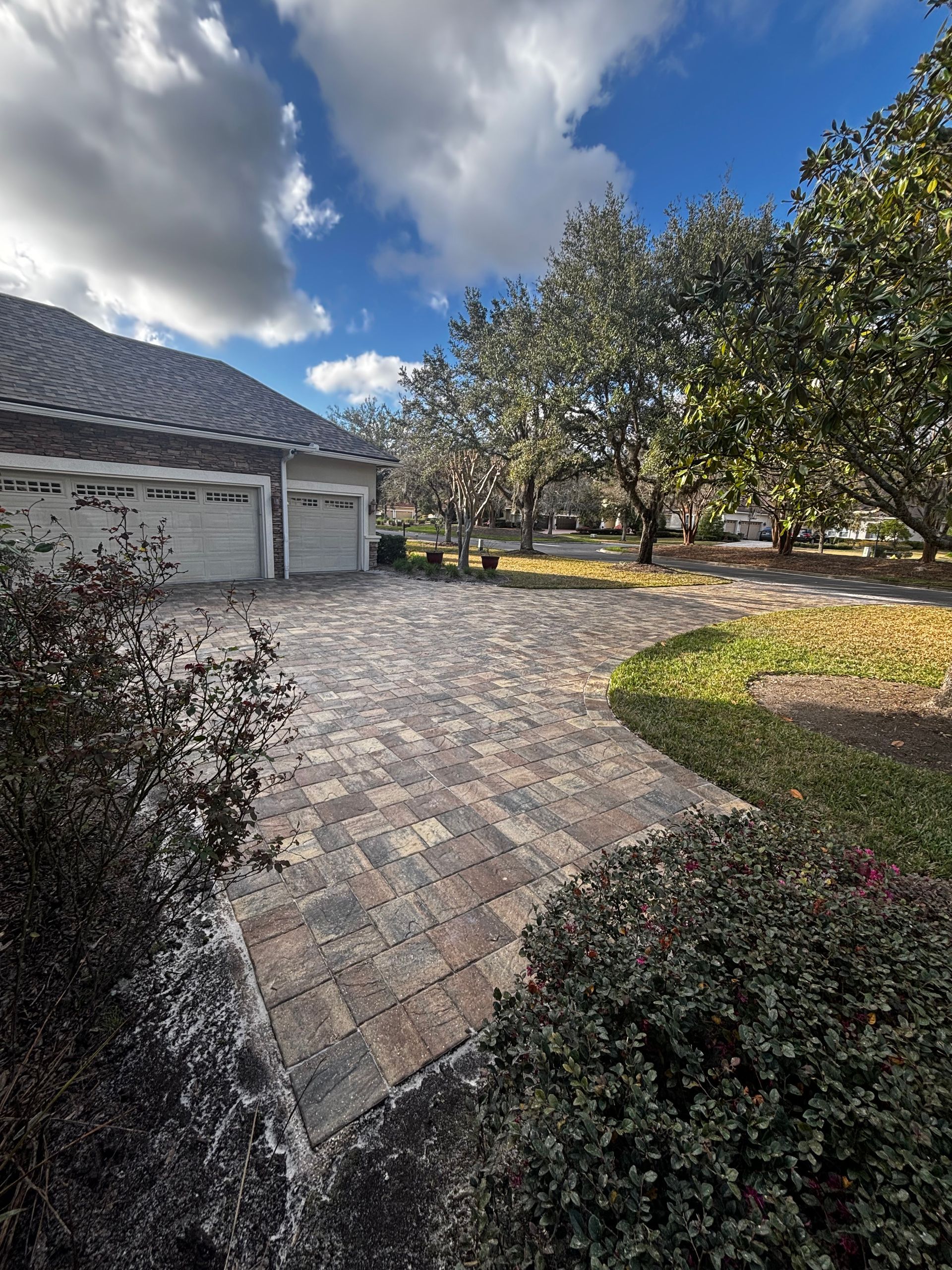 A paver driveway leads to a three-car garage in front of a house on a sunny day with trees and landscaping.
