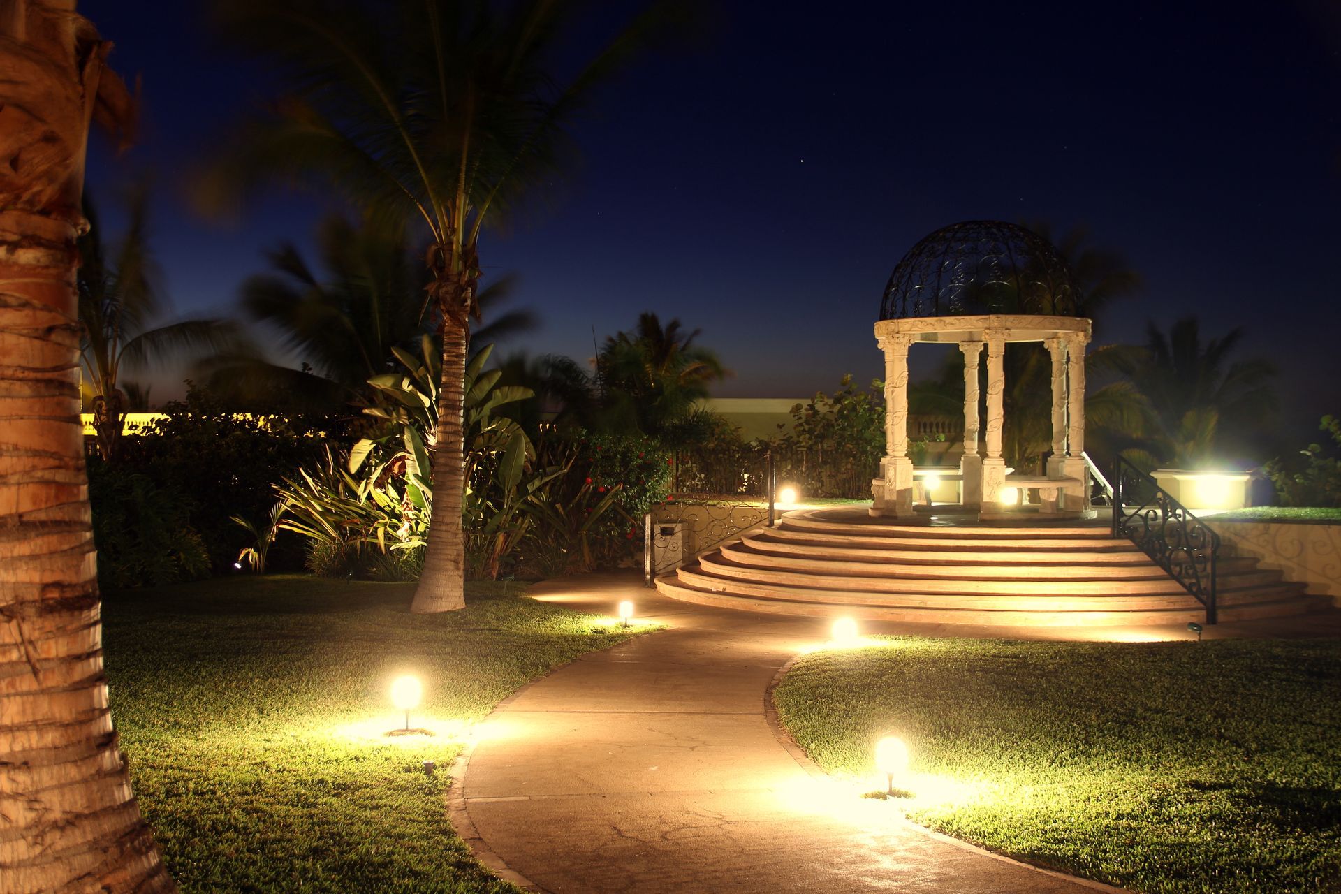 Elegant outdoor garden pathway at night, illuminated by soft ground lights, leading to a white stone