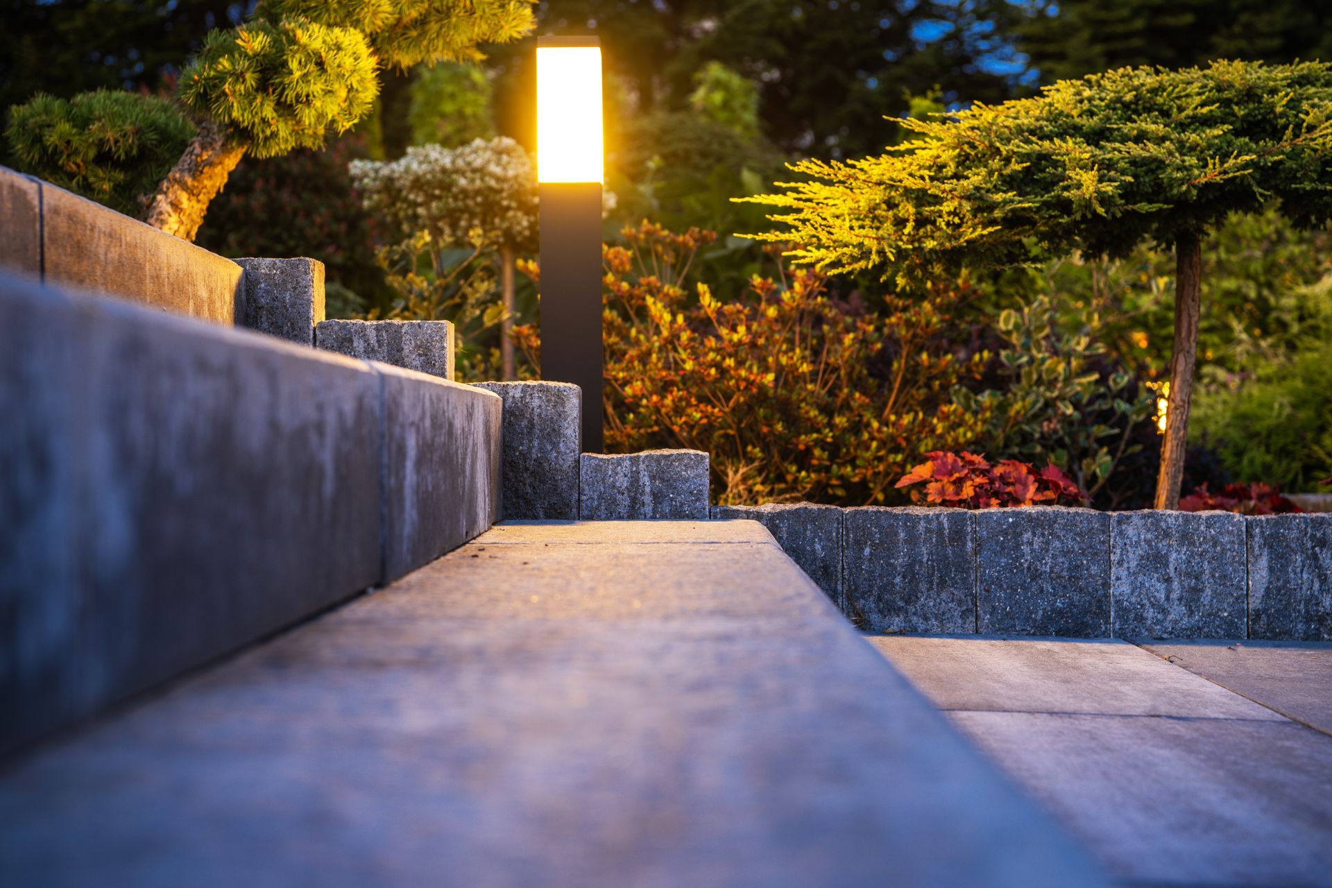 A bright, modern garden light stands beside stone steps in a lush, dimly lit garden at dusk.