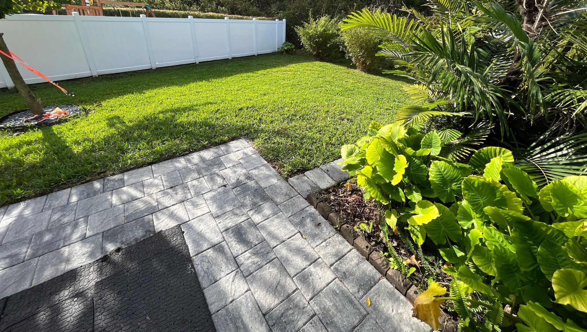 A backyard featuring a paved patio area, lush green grass, vibrant tropical plants.