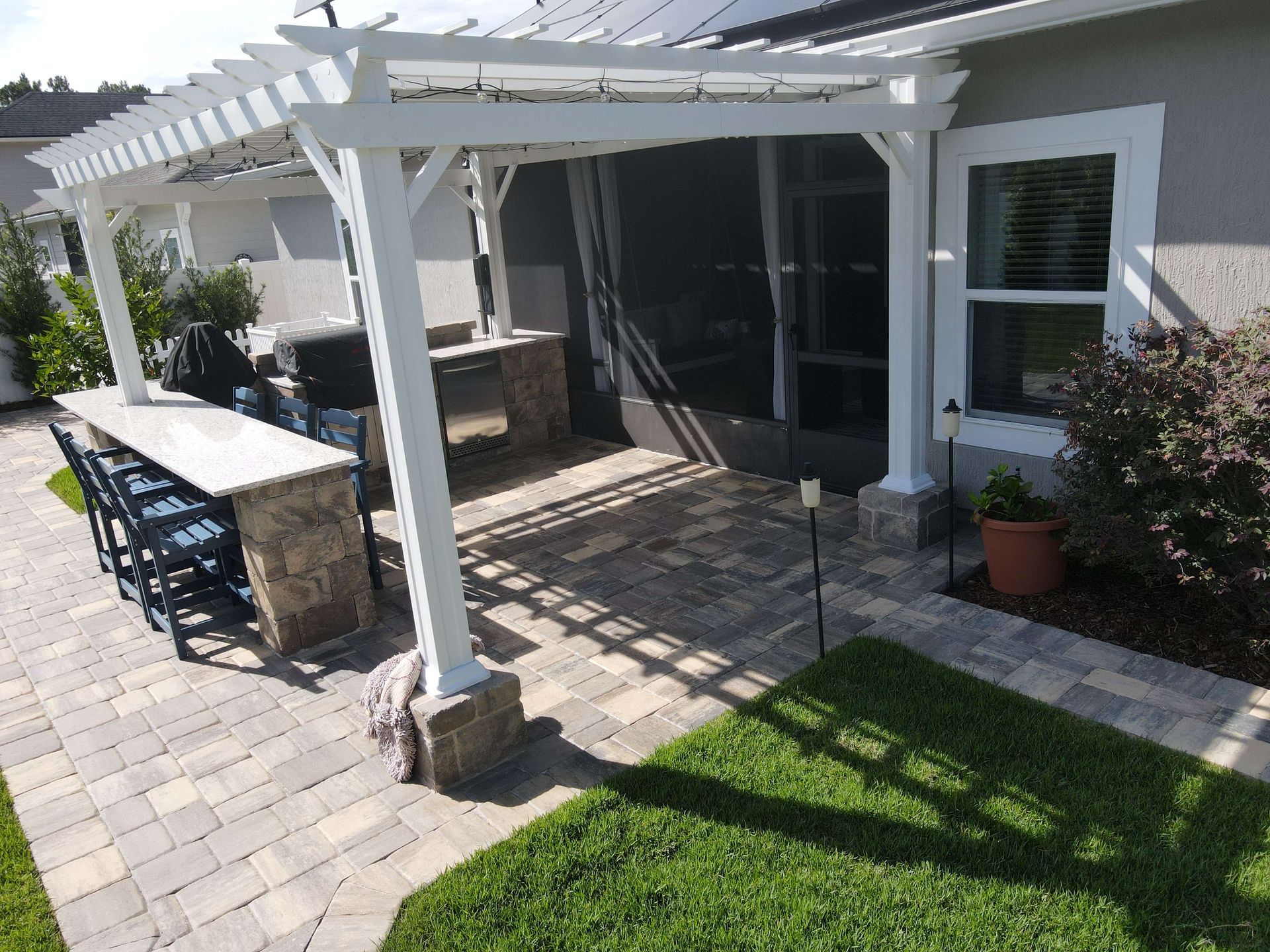 Outdoor patio with white pergola, stone pavers, built-in grill, and bar seating.