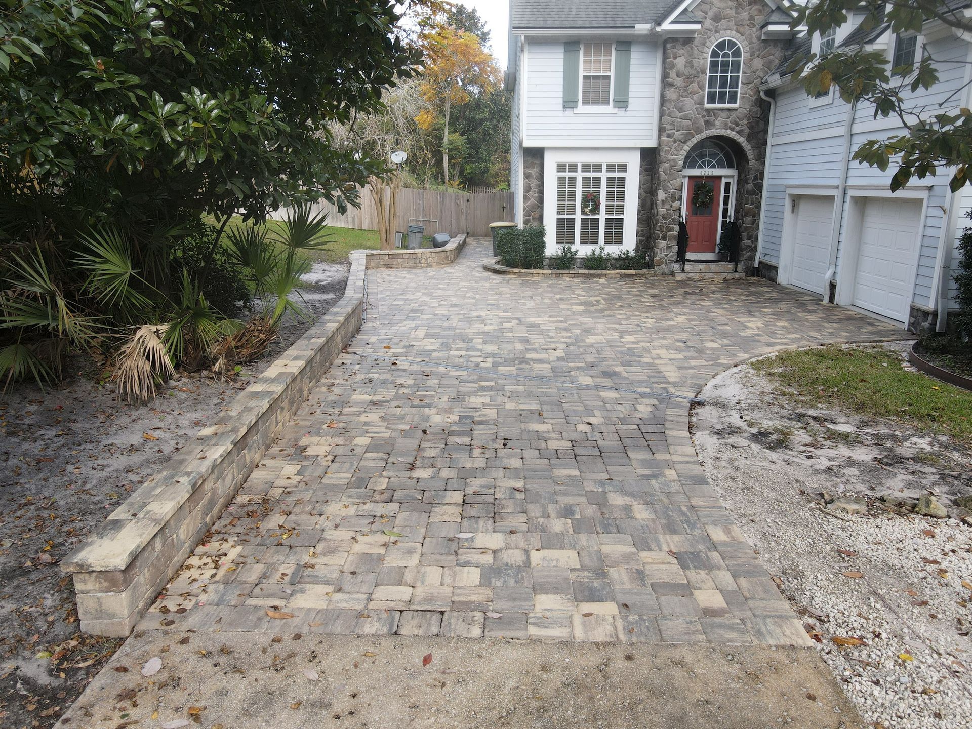 A paver driveway leads to a two-story house with a stone entryway, featuring a low retaining wall alongside garden beds.
