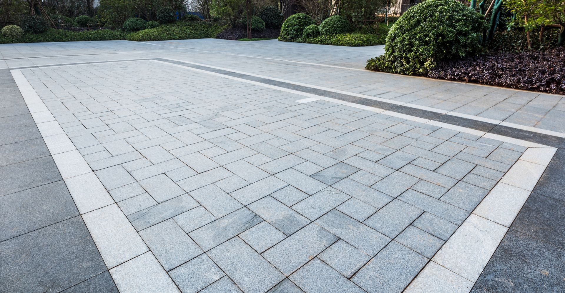 A light gray paved driveway with a rectangular border of lighter stone blocks, set against a garden area.