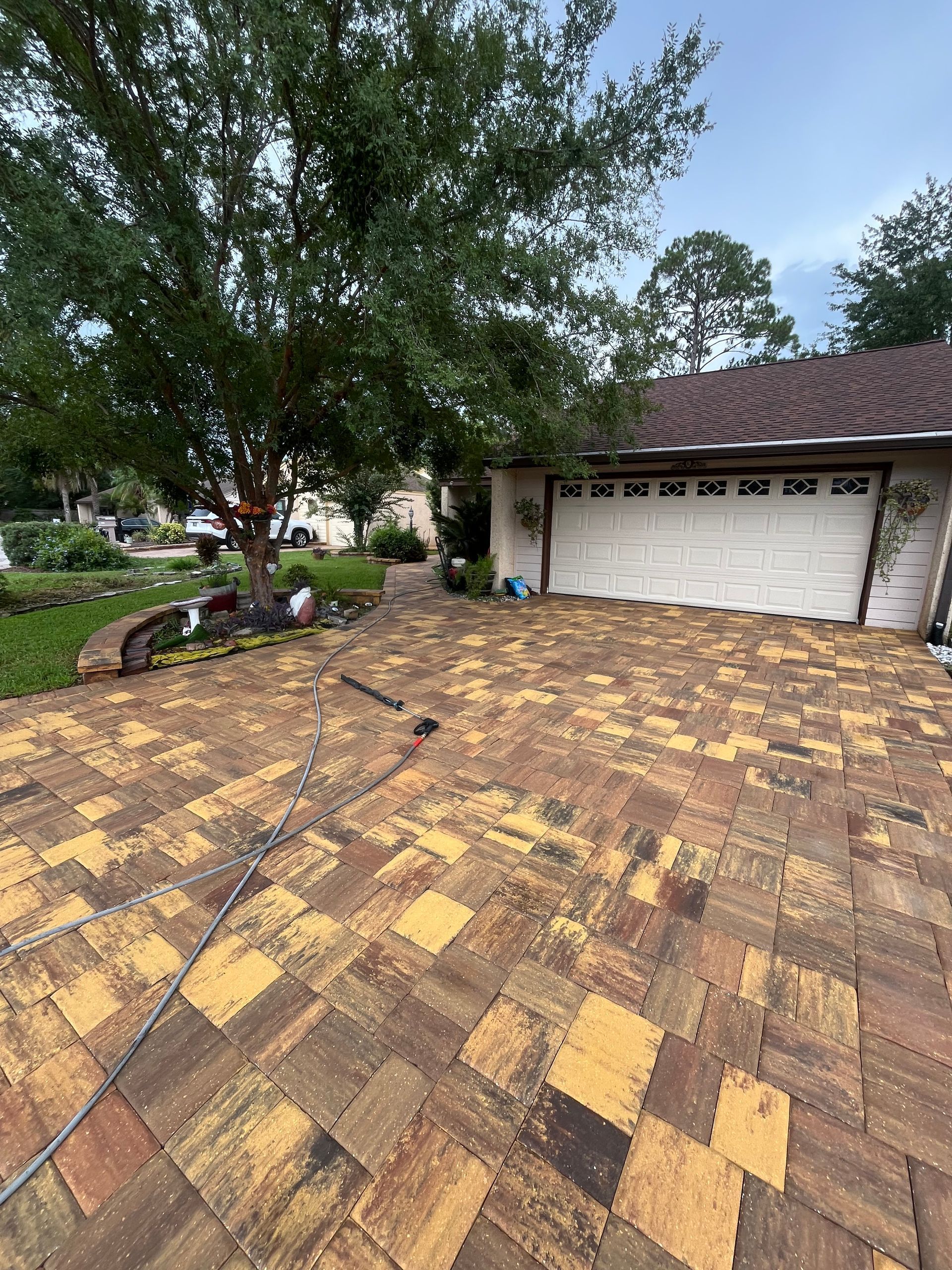 A driveway paved with rectangular stones in shades of brown, tan, and gold leading to a white garage under a large tree.