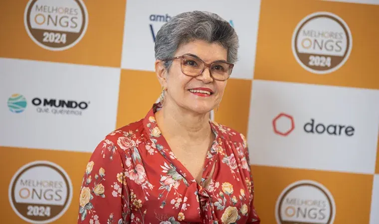 Uma mulher usando óculos e uma camisa florida está parada em frente a uma parede com logotipos de ongs.