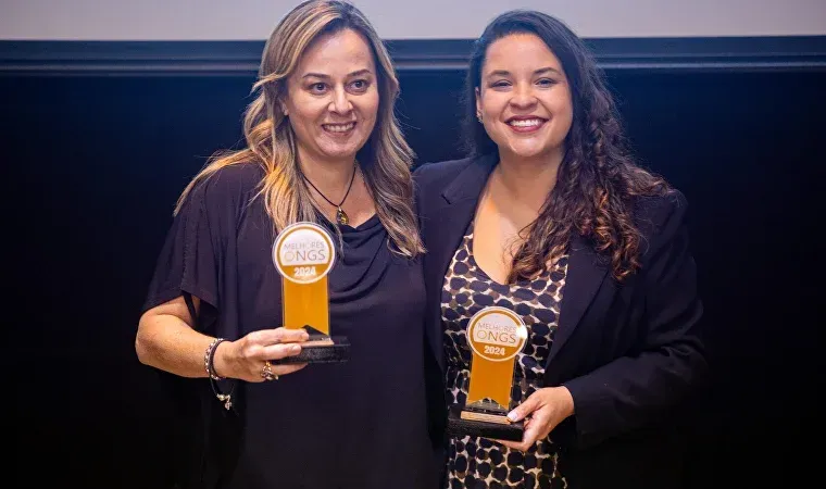 Duas mulheres estão de pé, uma ao lado da outra, segurando troféus.