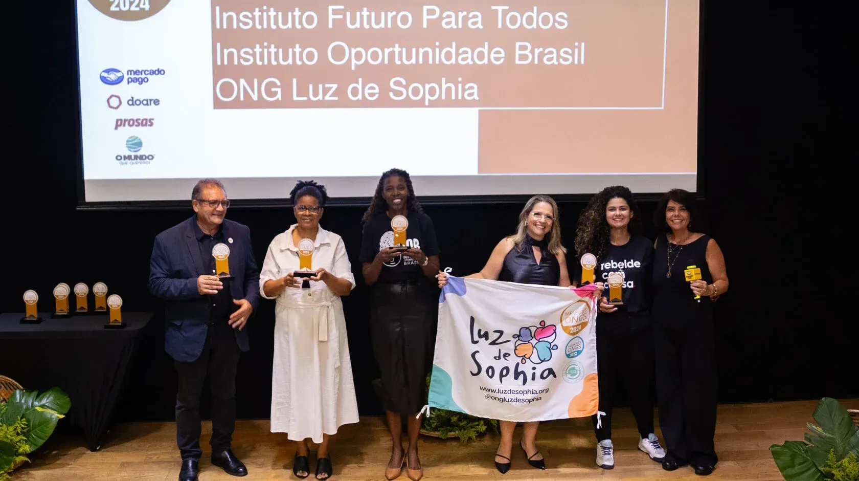 Um grupo de pessoas está em um palco segurando prêmios e uma bandeira.