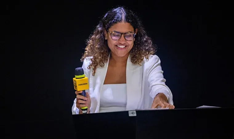 Uma mulher está sentada em uma mesa segurando um microfone e sorrindo.