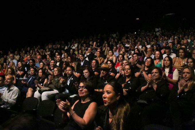 Uma grande multidão de pessoas está sentada em um teatro aplaudindo.