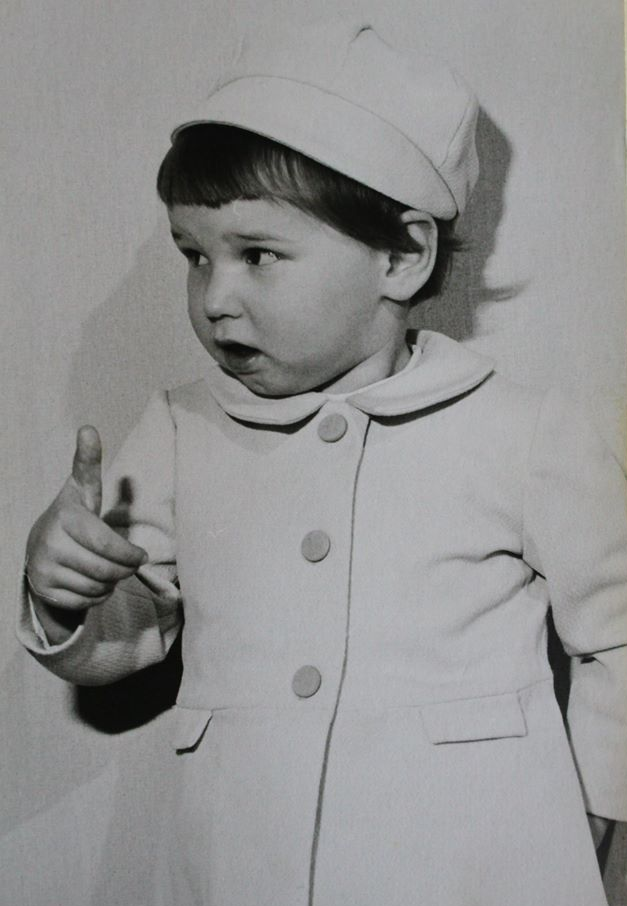 Jong kind in jas en hoed wijst met de vinger naar links. Zwart-witfoto.