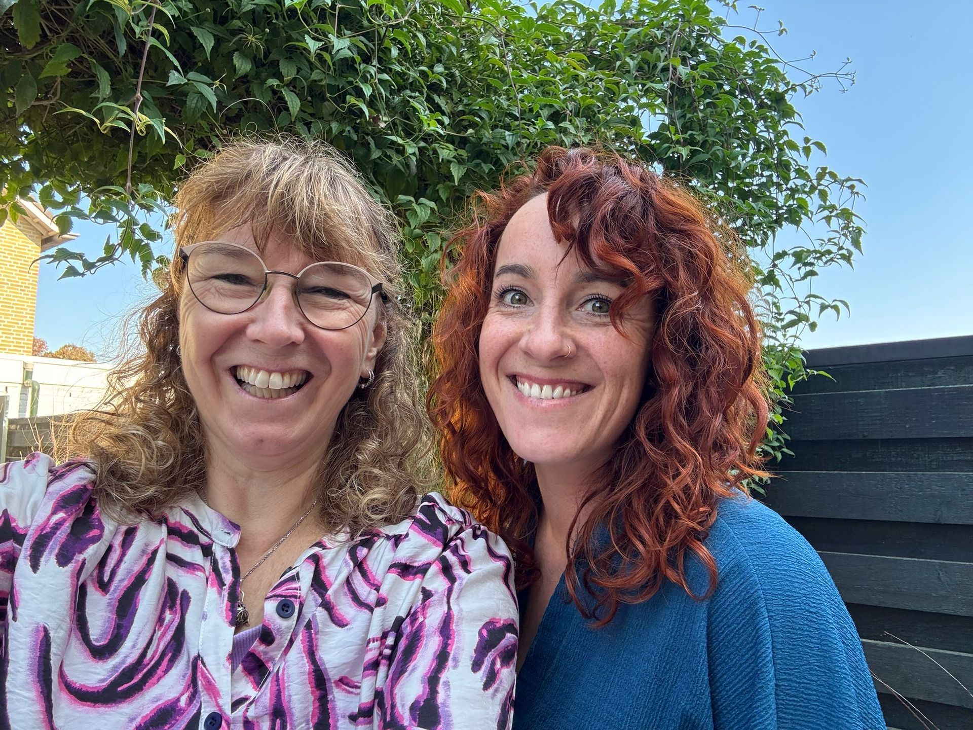 Twee lachende vrouwen staan ​​allebei voor een groen bladerdek.