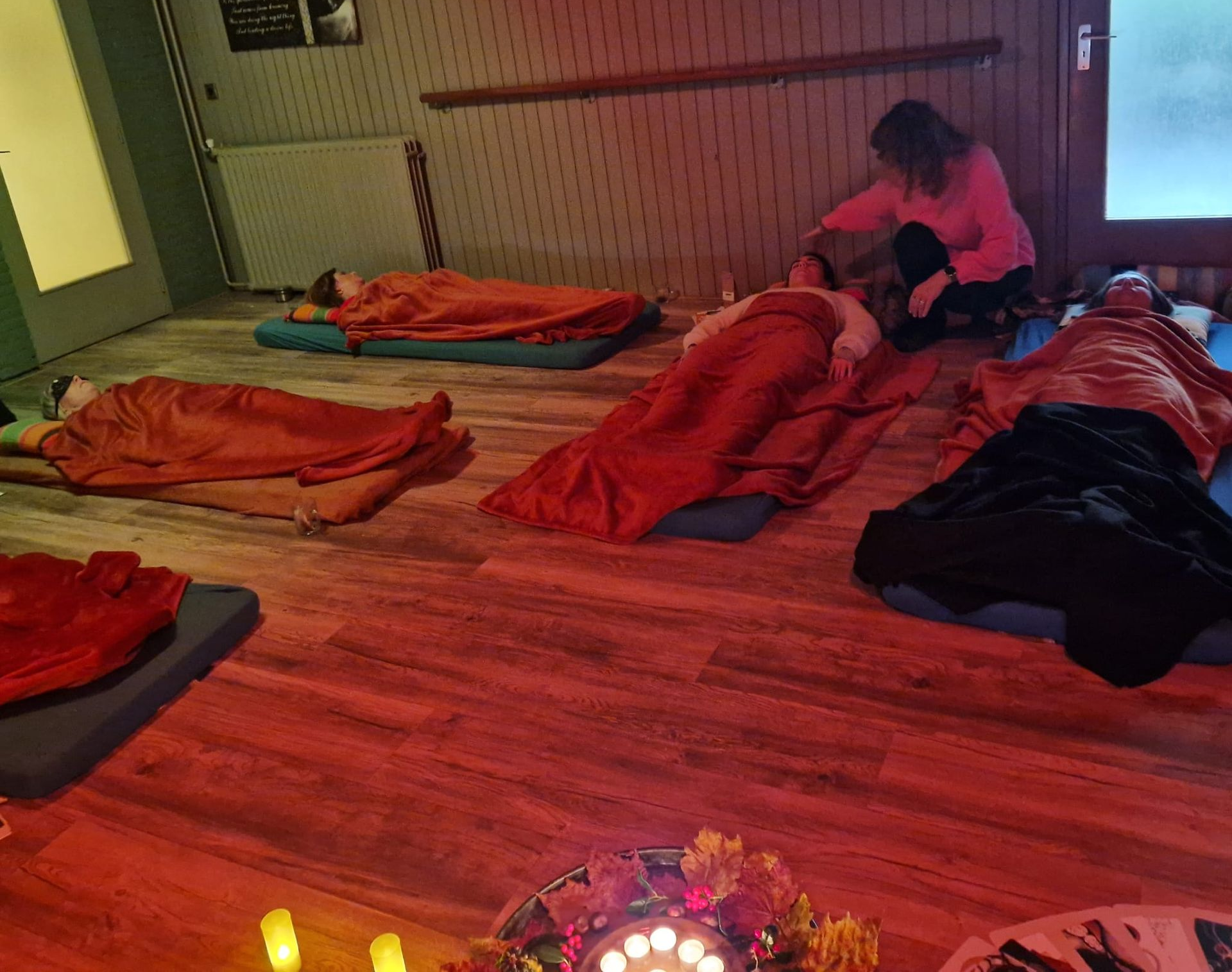 Mensen rusten op matten, bedekt met dekens, in een slecht verlichte kamer; een persoon biedt hulp.