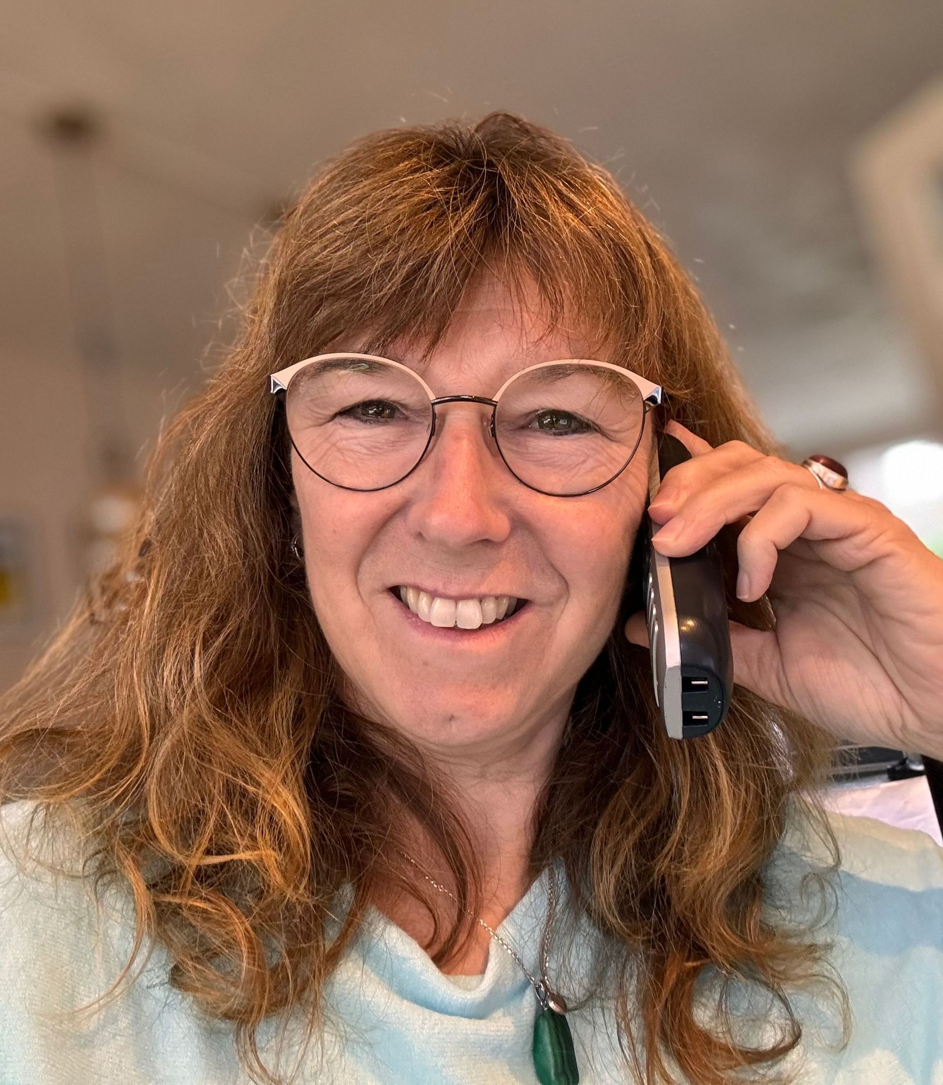 Vrouw met bril, telefonerend, glimlachend. Lichtblauw shirt, lang bruin haar.