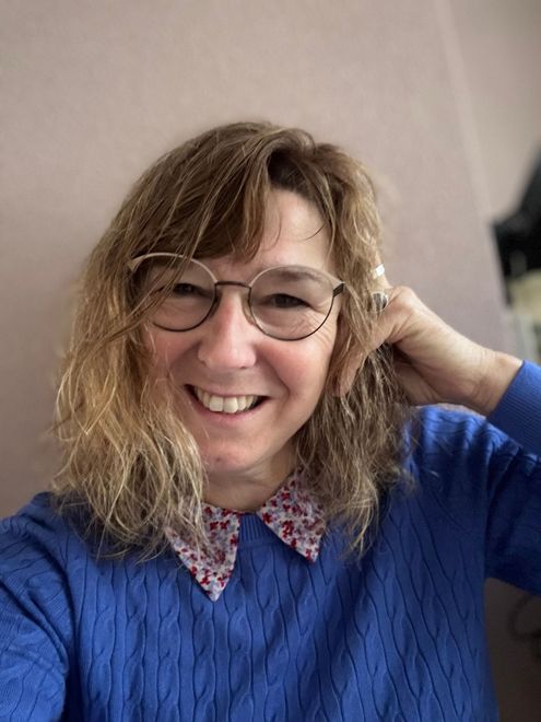 Vrouw met bril glimlacht, hand op haar hoofd, draagt ​​een blauwe trui met bloemenkraag.