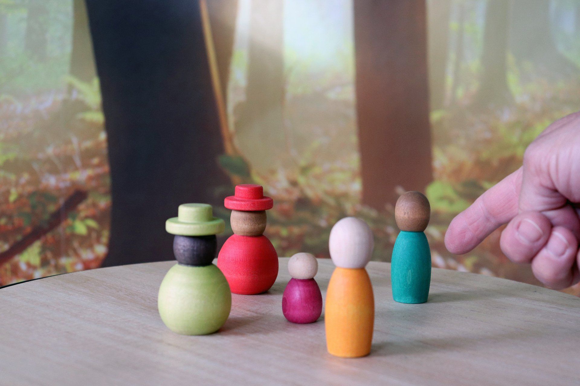 Houten poppetjes in een bosrijke omgeving; een hand wijst naar een van de poppetjes.