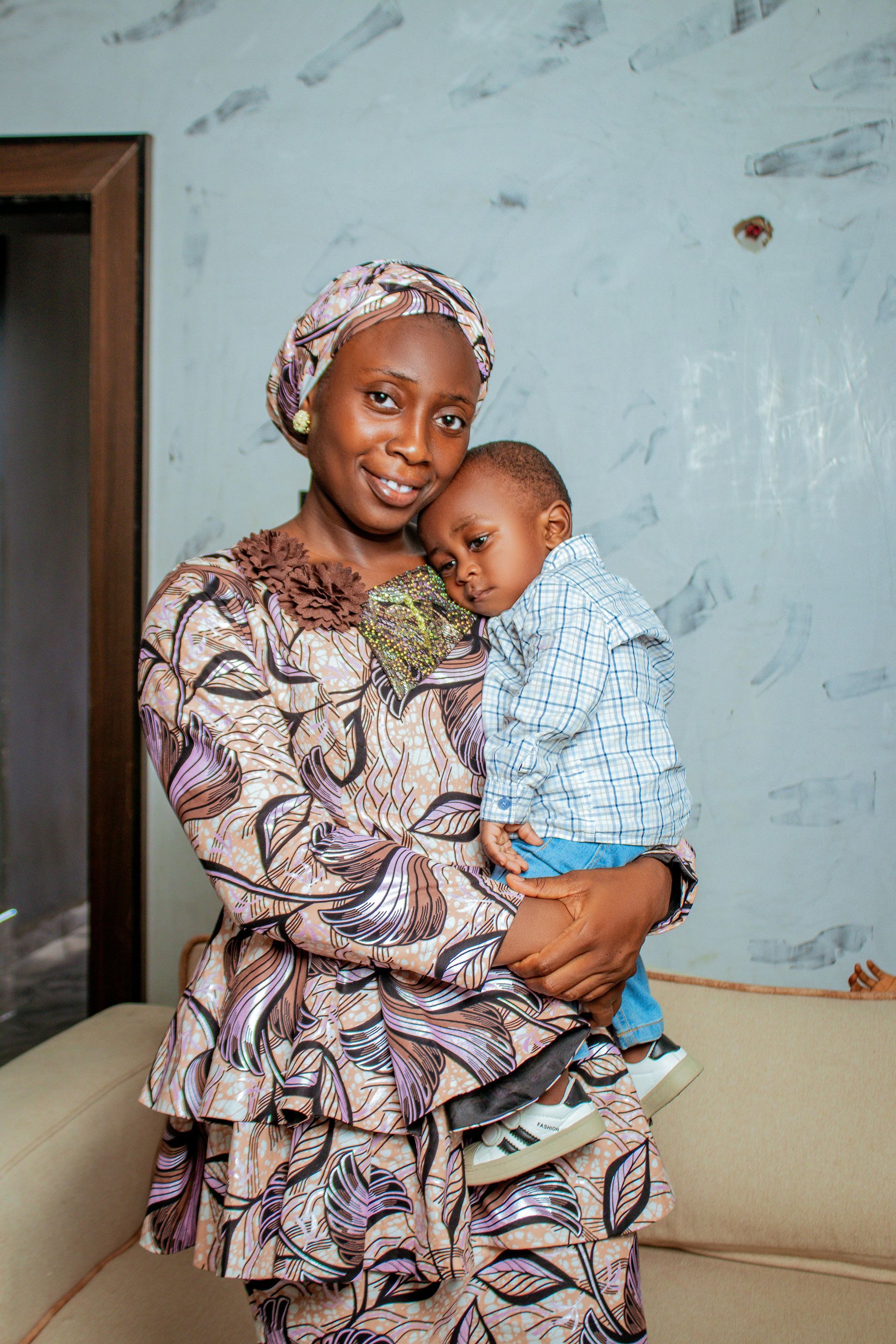 Woman in patterned outfit holding a sleeping baby. Both in front of a wall indoors.