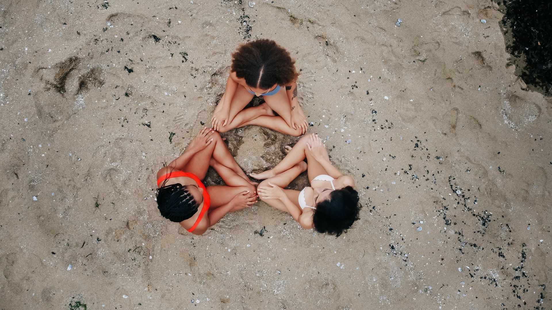 Drie vrouwen, in bikini, zitten in een driehoekige vorm op een zandstrand. Bovenaanzicht.