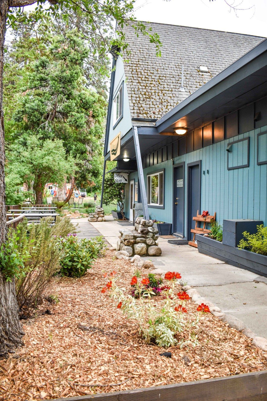 Blue cottage with flower beds and a stone walkway under trees