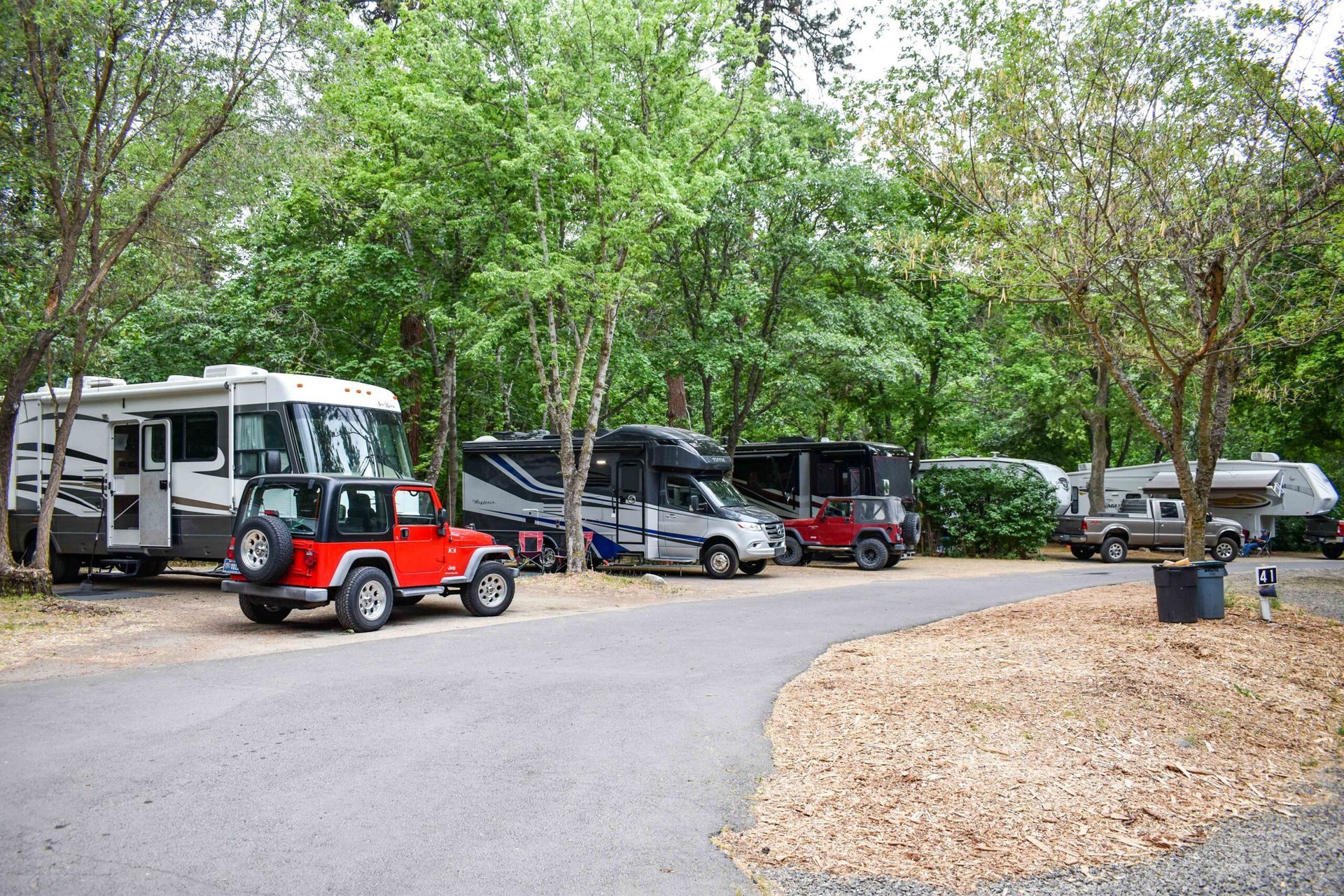 RV campsite with trees, parked trailers, and a red Jeep on a gravel road